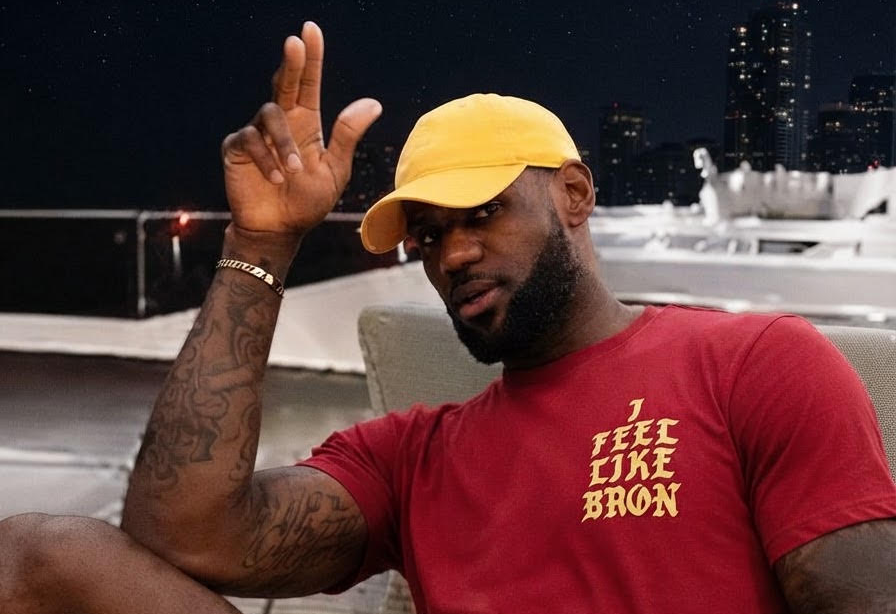 A man sitting outdoors at night wearing a red t-shirt with yellow text that reads 'I FEEL LIKE BRON' and yellow shorts, paired with a yellow cap. He is gesturing with his hand while holding cards.